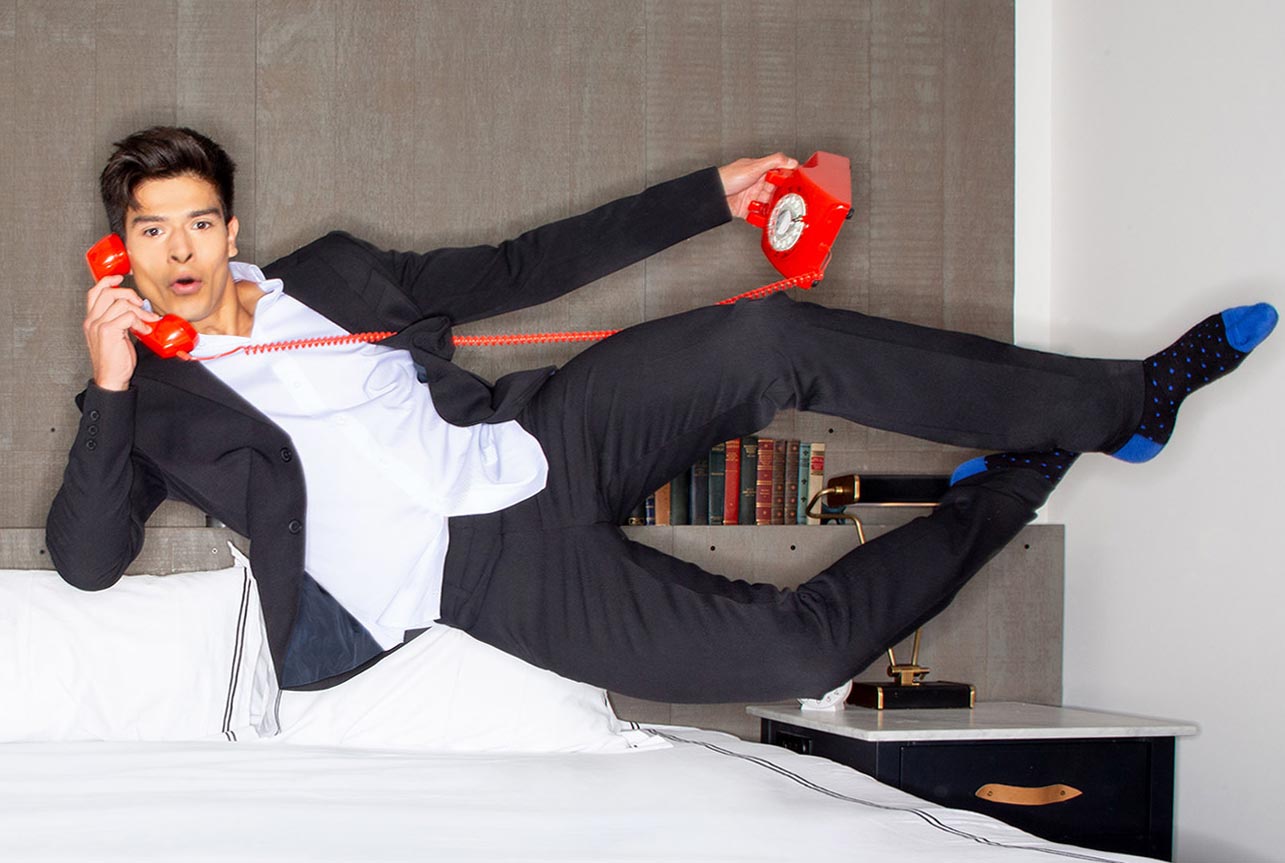 Modern Ambition male model flying onto a bed wearing a suit and talking on a red retro telephone