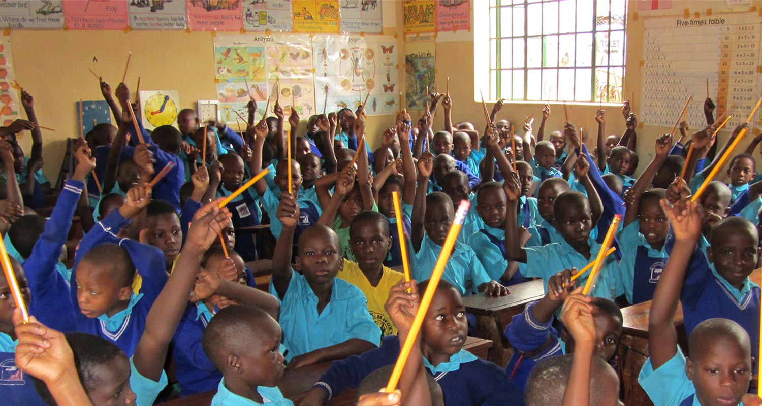 School children in a classroom with raised hands, at Kamwokya Primary School