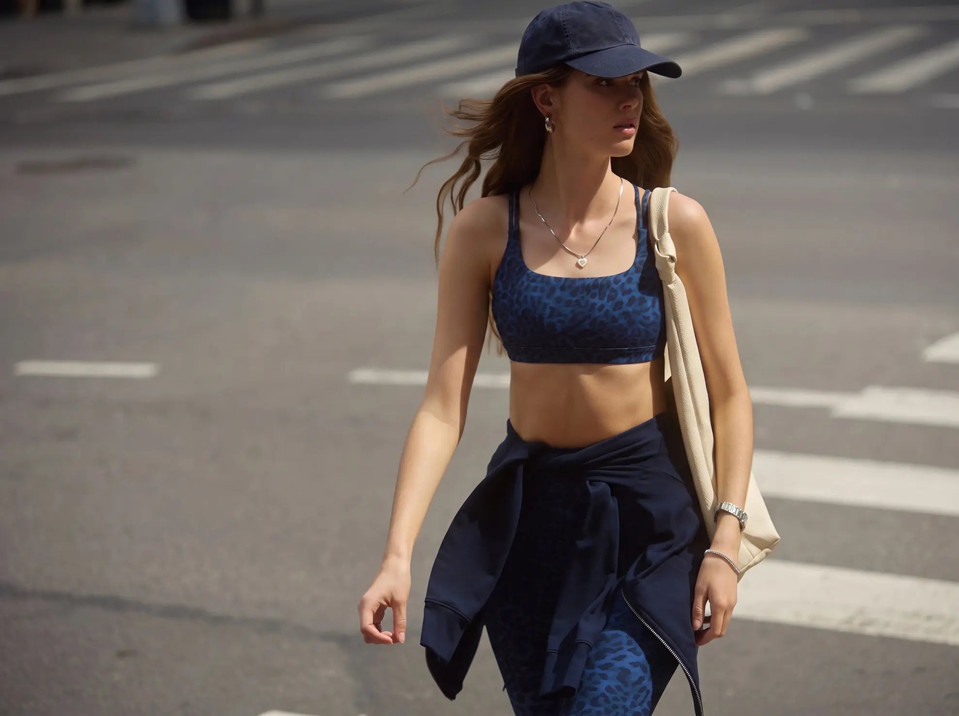 A woman walking on a city street wearing a navy blue leopard-print sports bra and matching leggings, a navy baseball cap, and carrying a beige shoulder bag.