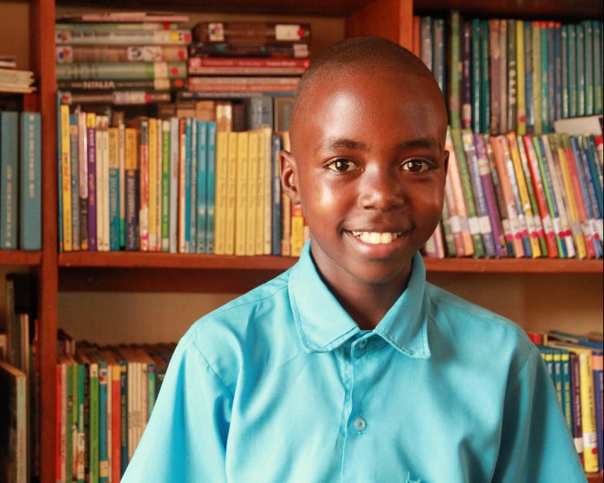Young boy in a blue shirt smiling in front of a bookshelf.