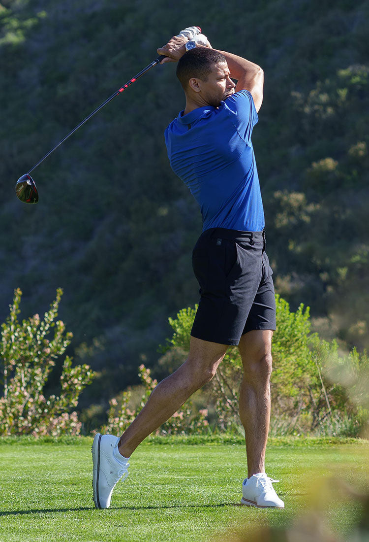 MPG male model on a hillside golf course, wearing a blue golf shirt and black shorts, tall image